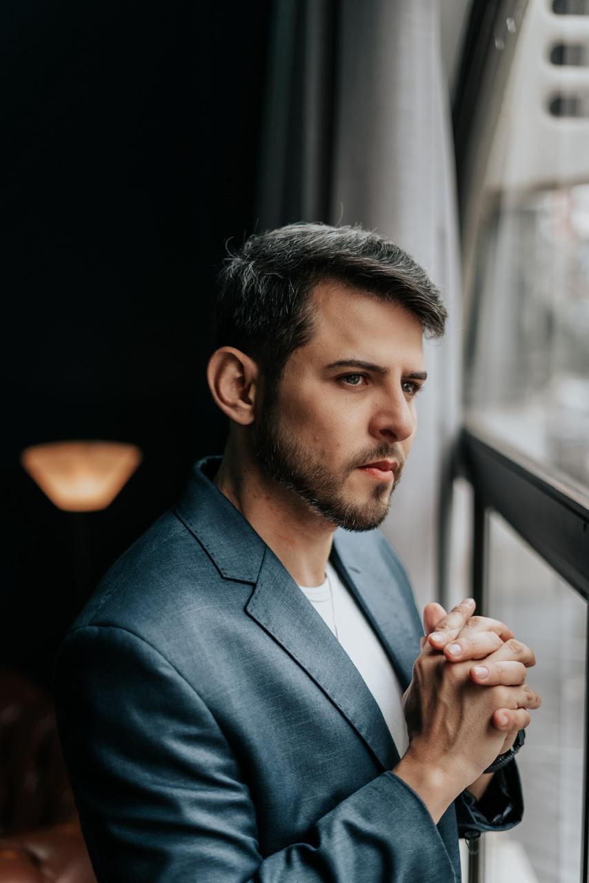 Diego pensativo junto à janela, refletindo sobre a decisão de investir
